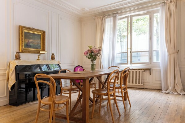 A dining room with a piano, wooden chairs, a table, and a window with curtains.