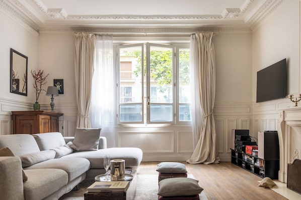Un salon comprenant un canapé, une table basse, une cheminée, un téléviseur et une fenêtre avec des rideaux.