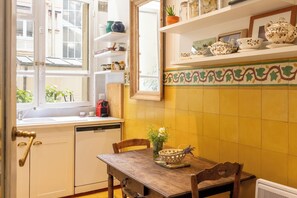 A kitchen with a window, a sink, a dishwasher, a wooden table with a vase, and shelves with kitchenware.