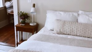 Ein ordentlich bezogenes Bett mit weißen Leinen und einer gesteppten Decke, ein Nachttisch mit Lampe und einer Blumenvase sowie ein Holzboden.