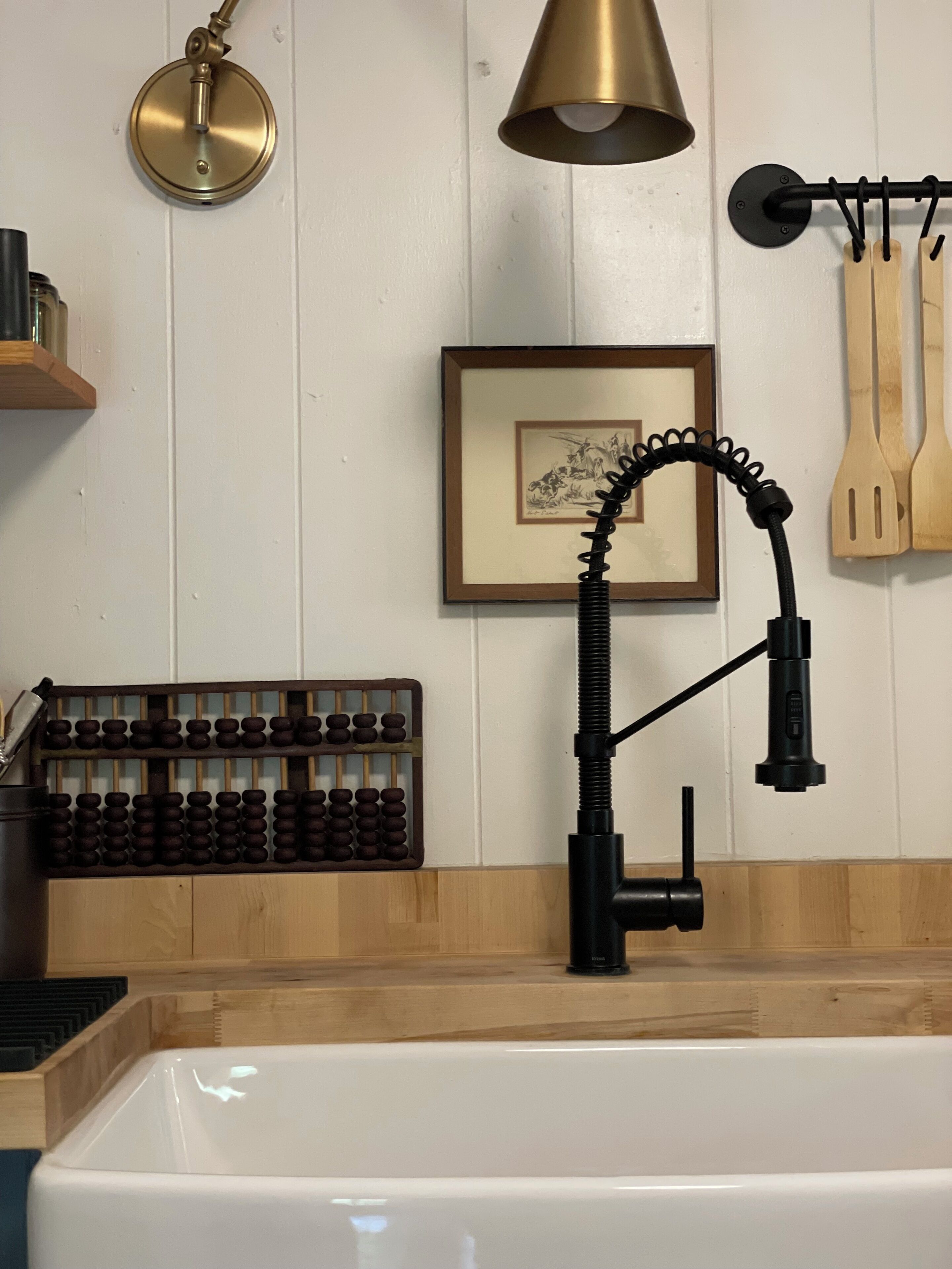 Une cuisine avec un évier blanc, un robinet noir, des ustensiles de cuisine en bois accrochés au mur et un tableau encadré.