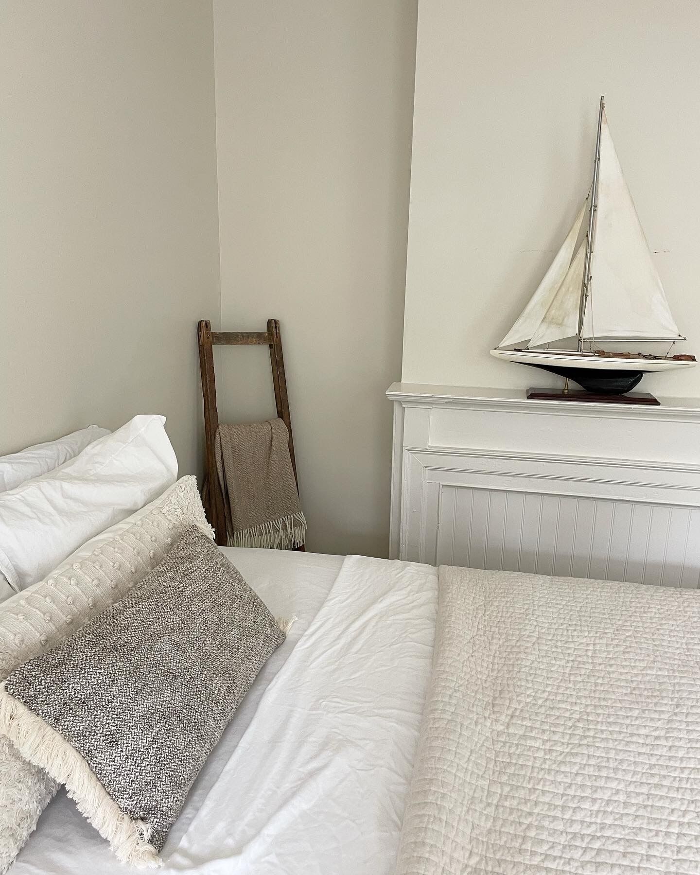 A bedroom with a bed, pillows, a wooden chair, and a sailboat model on a shelf.