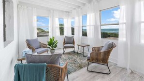 Una habitación luminosa con ventanas amplias, mobiliario de mimbre, una mesita y una planta en maceta.