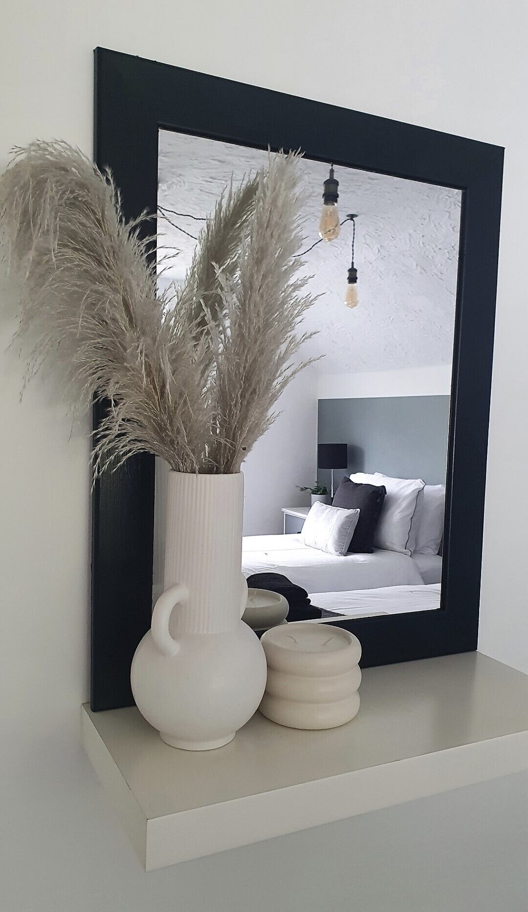 A bedroom with a bed, bedside table, lamp, and a mirror with a black frame.