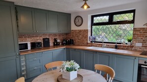 A kitchen with green cabinets, a wooden table with chairs, a clock on the wall, and a window with a view of greenery. - Green Cottage (Bangor)