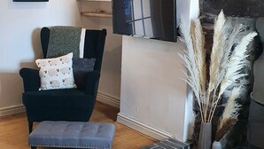 A modern living room with a stone fireplace, a black leather sofa, a flat-screen TV, and a decorative vase with pampas grass. - Green Cottage (Bangor)