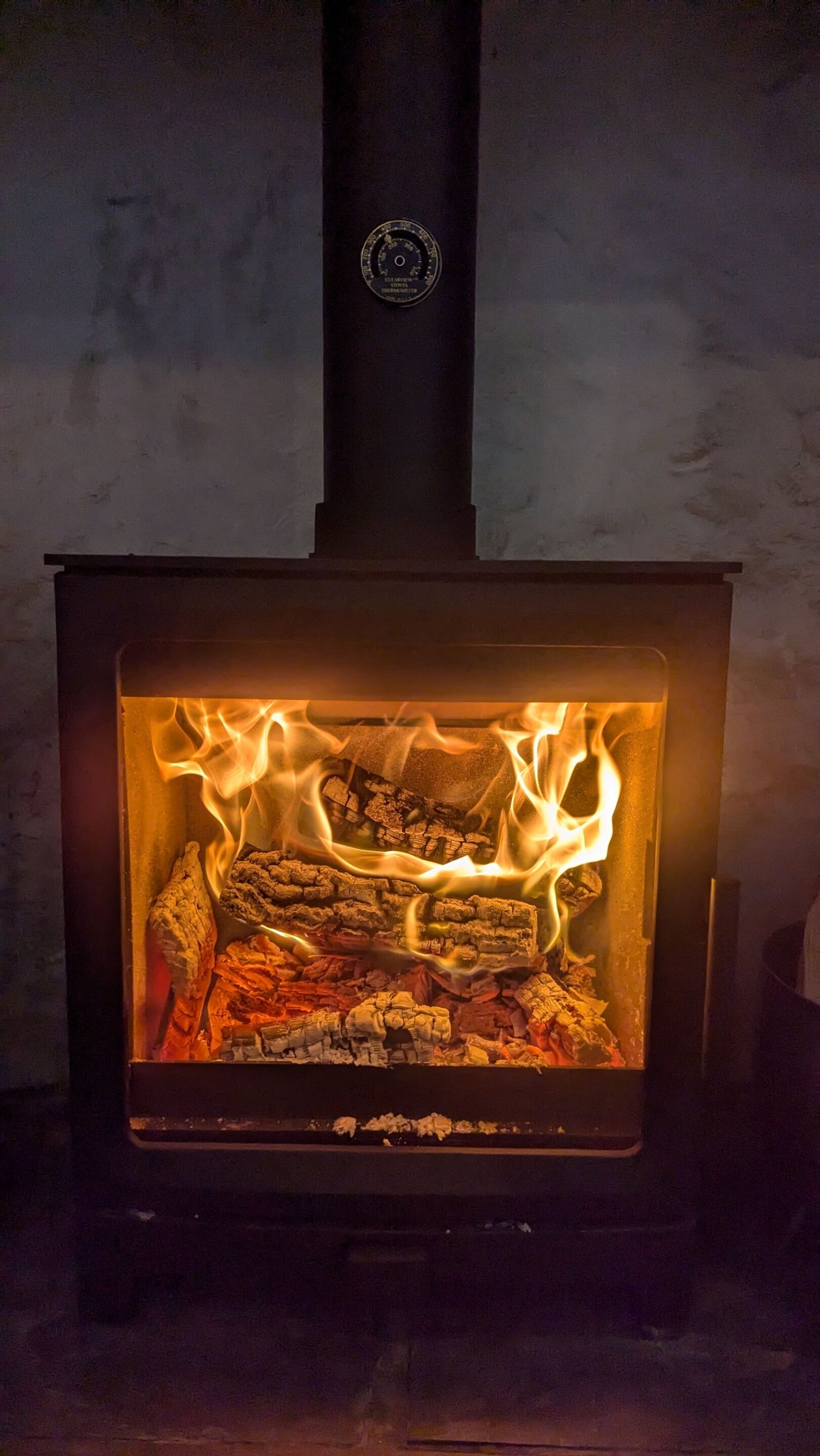 A wood-burning stove with visible flames, a black chimney, and a circular logo on the chimney.