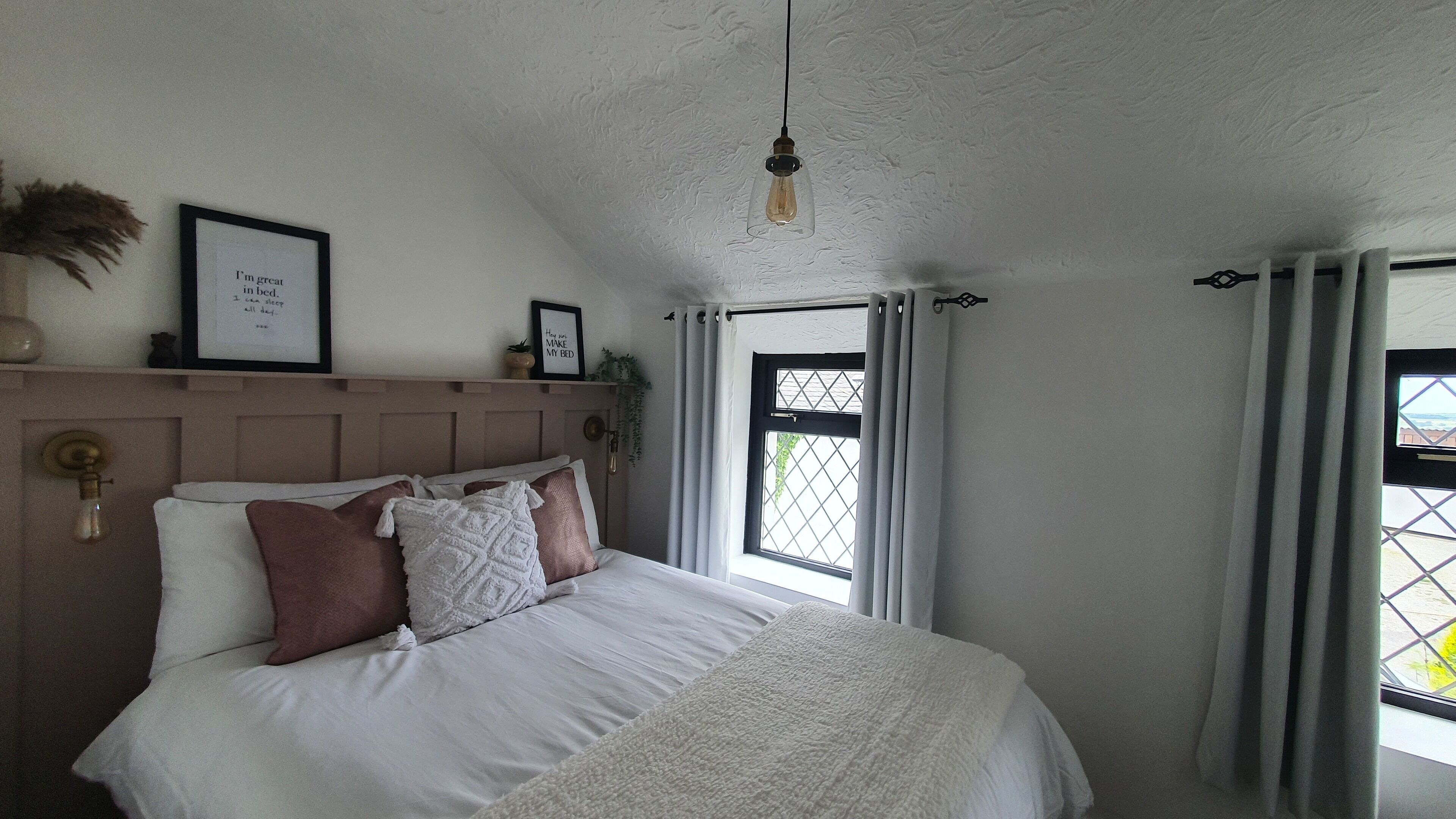 A bedroom with a bed, a hanging light fixture, a window with curtains, and a wall-mounted shelf with decorative items.