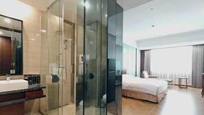 Una habitación de hotel moderna con ducha de vidrio, una cama grande, una mesita de noche y vistas de la ciudad a través de amplios ventanales.