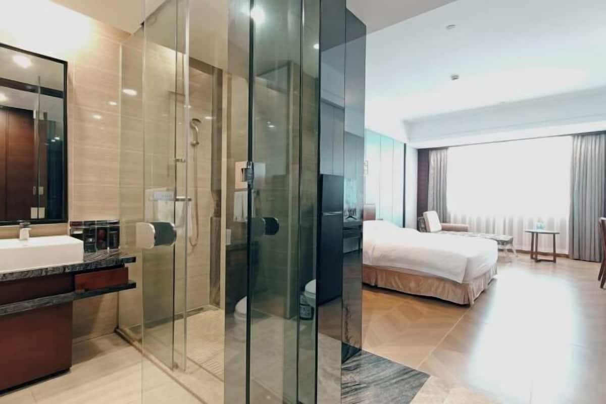 Una habitación de hotel moderna con ducha de vidrio, una cama grande, una mesita de noche y vistas de la ciudad a través de amplios ventanales.