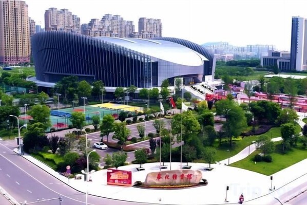A modern stadium with a curved roof, surrounded by greenery and a sports field.