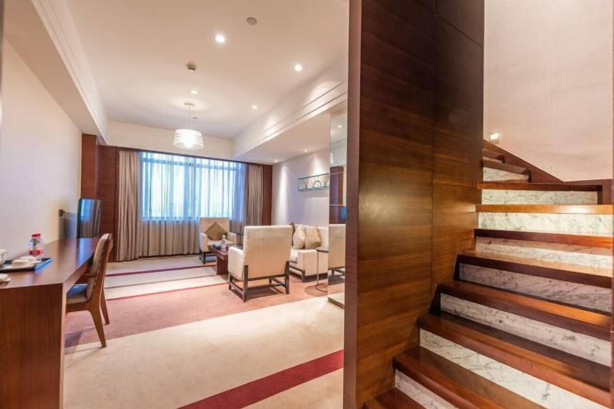 Habitación de hotel con escalera de madera, zona de estar con sillas, mesa de comedor y un gran ventanal con cortinas.