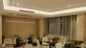 A hotel room with a dining area featuring multiple tables and chairs, a large window with curtains, and a tiled wall.