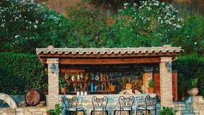 A rustic outdoor bar with stone walls, metal chairs, and a variety of bottles and glasses. - Traditional apartement with pool for relaxation moments (Trikorfo xalkeias)