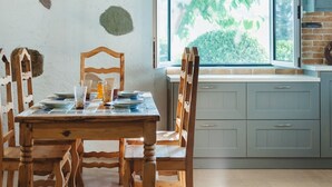 A dining area with a wooden table and chairs, a window with a view of greenery, and a kitchen with grey cabinets. - Traditional apartement with pool for relaxation moments (Trikorfo xalkeias)