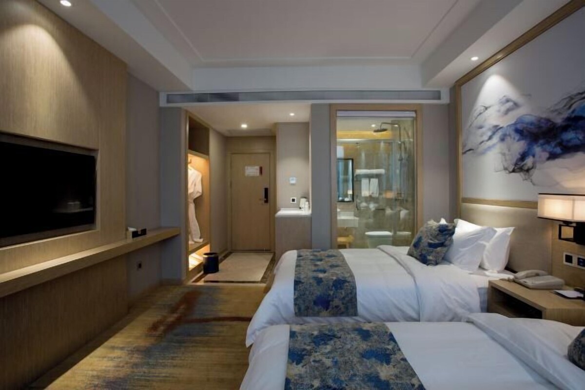 A hotel room with two beds, a TV, a bathroom with a glass door, and a painting on the wall.