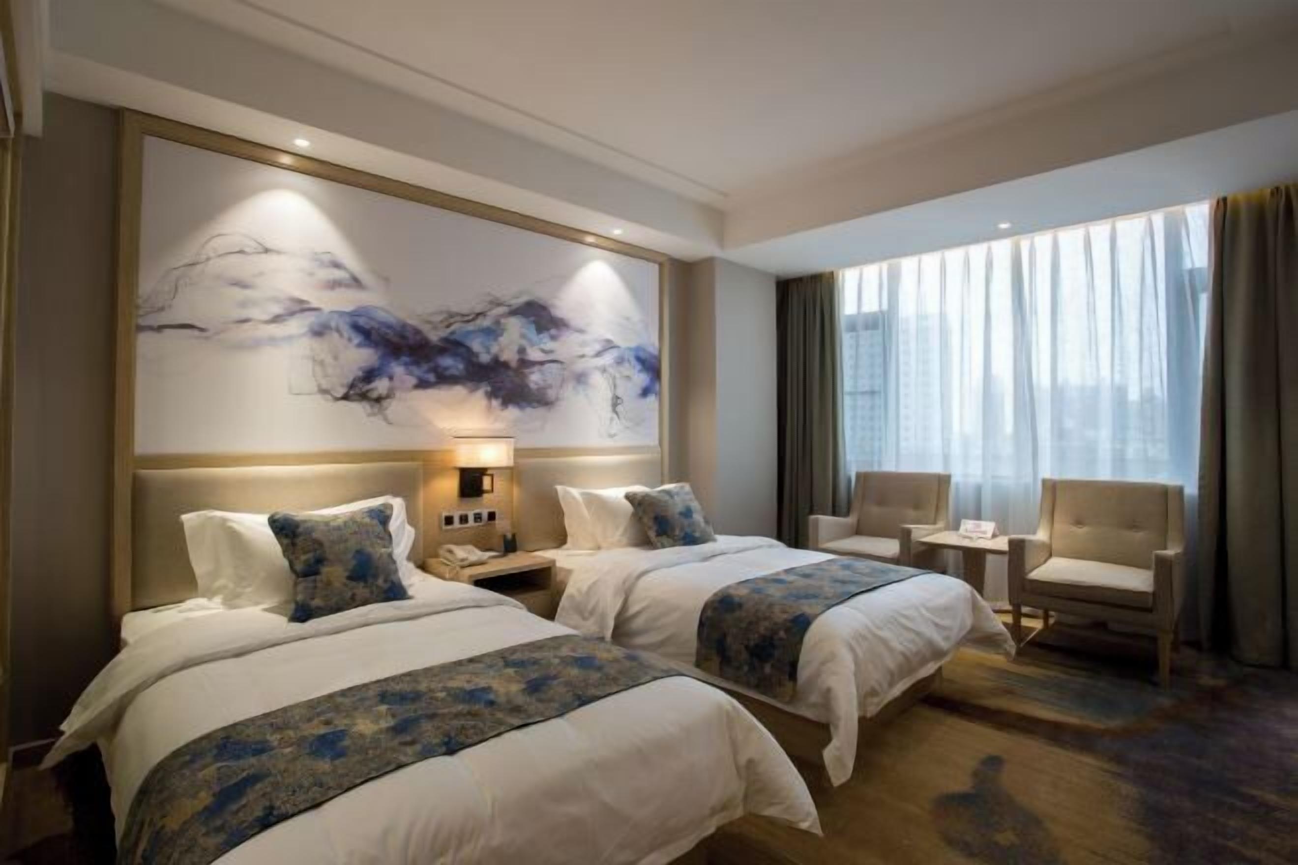 Camera d'albergo con due letti, un grande quadro astratto appeso alla parete, un comodino con una lampada, un tavolino, due sedie e un'ampia finestra con tende.