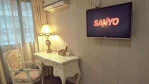 Una habitación con un escritorio blanco, una silla, una lámpara y un televisor que muestra 'SANYO' en la pared.
