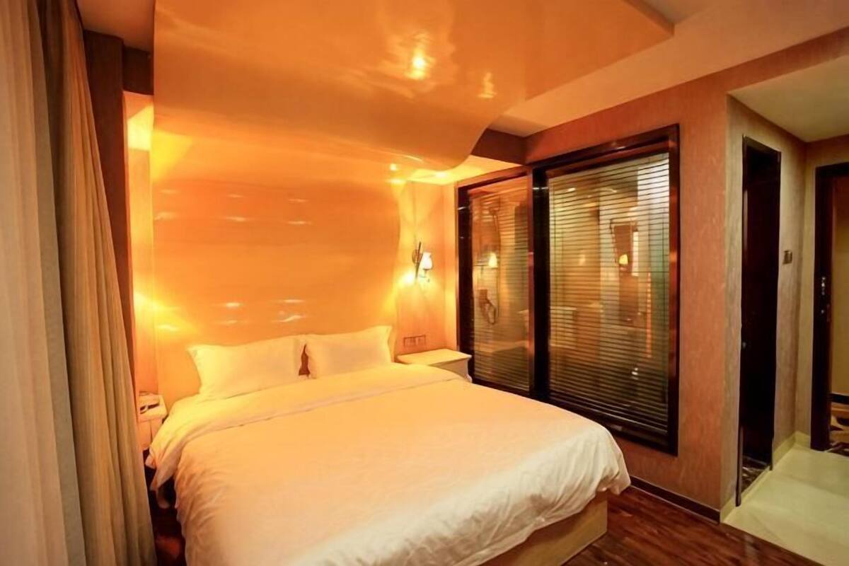 Une chambre d’hôtel avec un grand lit, une fenêtre avec des stores, une table de chevet et une porte.