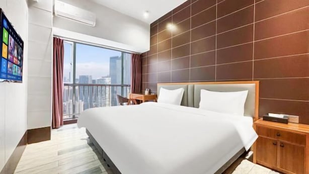A modern hotel room with a large bed, a wooden headboard, a nightstand, and a view of the cityscape. - Green Leaf Inn (Dongguan)