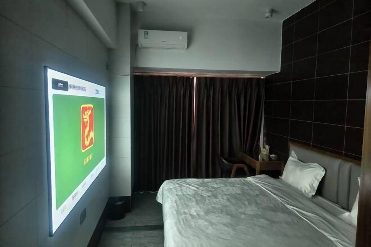 Habitación de hotel con cama, escritorio, silla y una gran pantalla digital que muestra un logotipo.