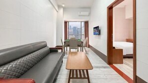 A hotel room with a sofa, a dining table, a TV, and a view of the city. - Green Leaf Inn (Dongguan)