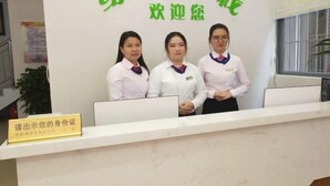 A reception desk with three staff members standing behind it. There is a sign above the desk with green text, and a sign below the desk instructing to show identification. - Green Leaf Inn (Dongguan)