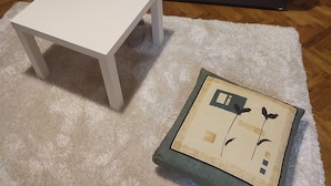 Une table blanche et un objet décoratif sont posés sur un tapis blanc dans une pièce où se trouve un canapé gris foncé.