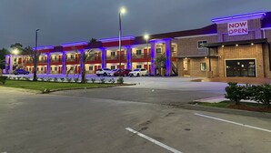 Un hôtel à plusieurs étages, reconnaissable à son panneau « OUVERT MAINTENANT », avec une façade en briques et un parking où sont stationnées plusieurs voitures.