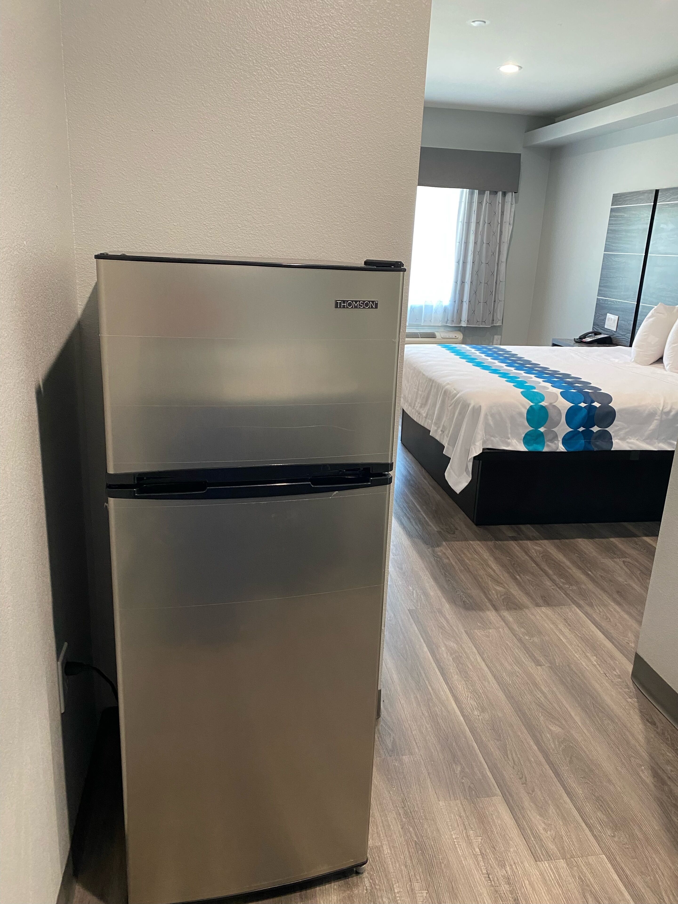 A modern hotel room with a built-in Thomson refrigerator, a neatly made bed with a blue and white patterned bedspread, and a wooden floor.