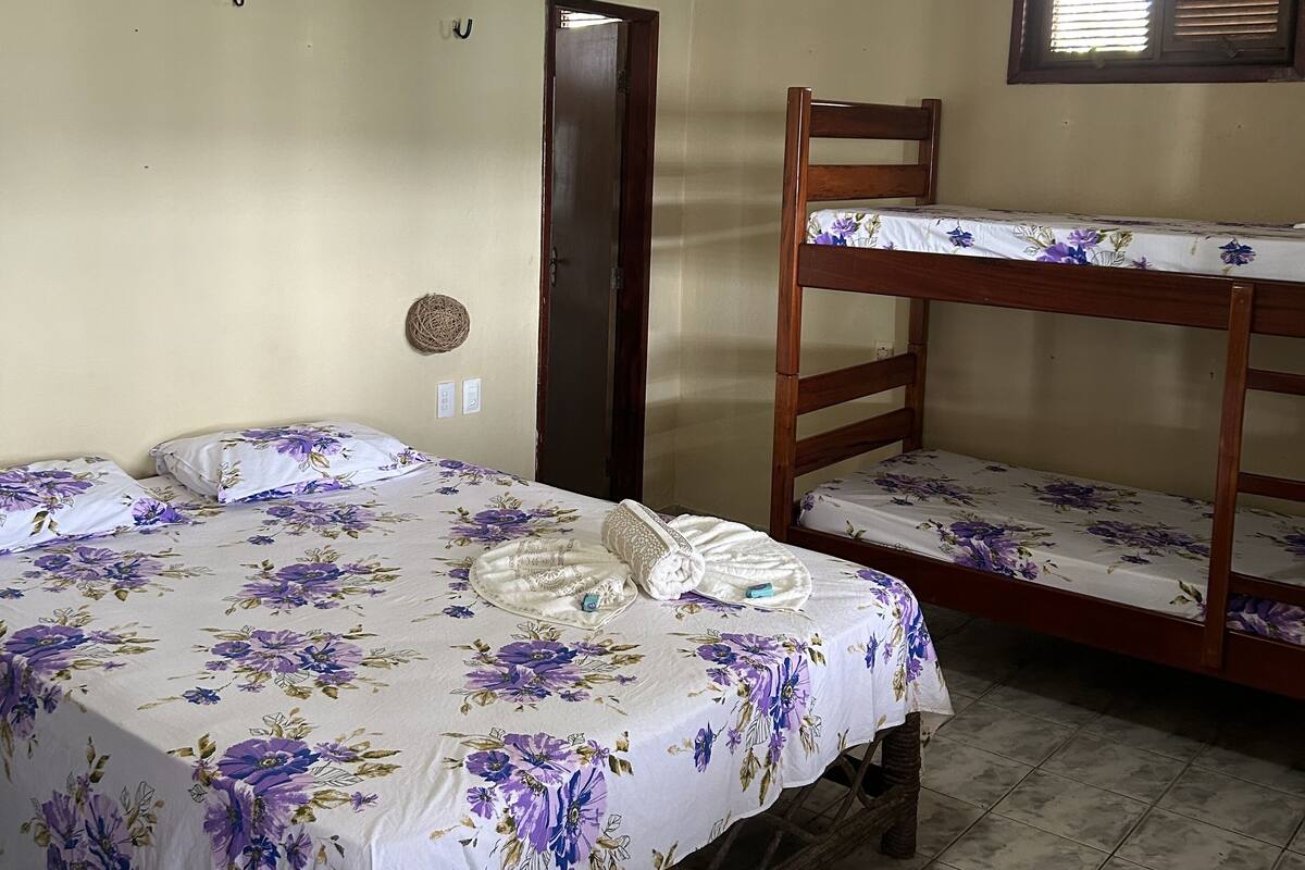 Quarto com cama de solteiro, uma cama individual com roupa de cama florida, ventilador de teto, janela com persianas e uma porta.