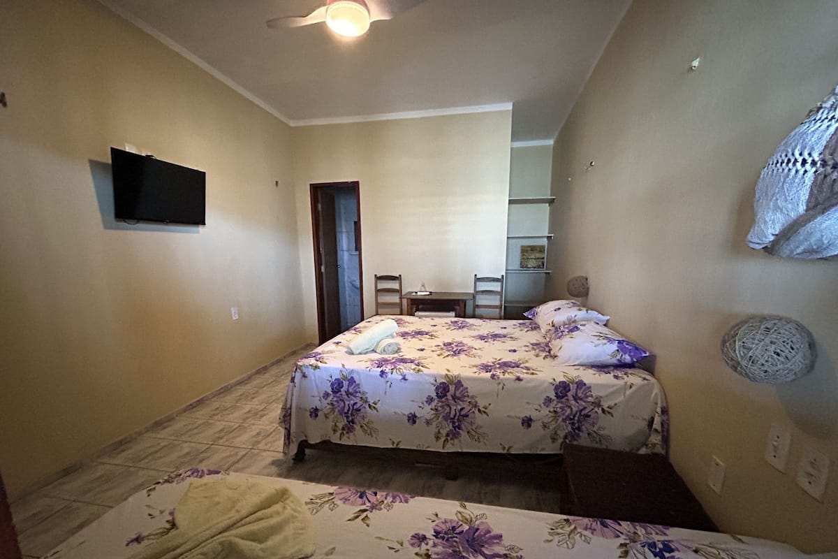 Quarto com cama, uma cadeira, escrivaninha e televisão na parede.