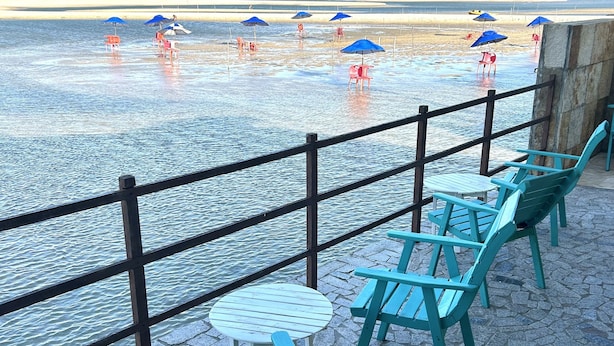 Terraza frente al agua con sillas y mesas, barandilla y sombrillas.