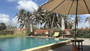 Un área de piscina con sillones de descanso y sombrillas, rodeada de palmeras y un muro de piedra.