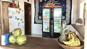 Zona de bar con un refrigerador lleno de bebidas, una estantería de madera con botellas, y un cuenco de plátanos y cocos en la encimera.