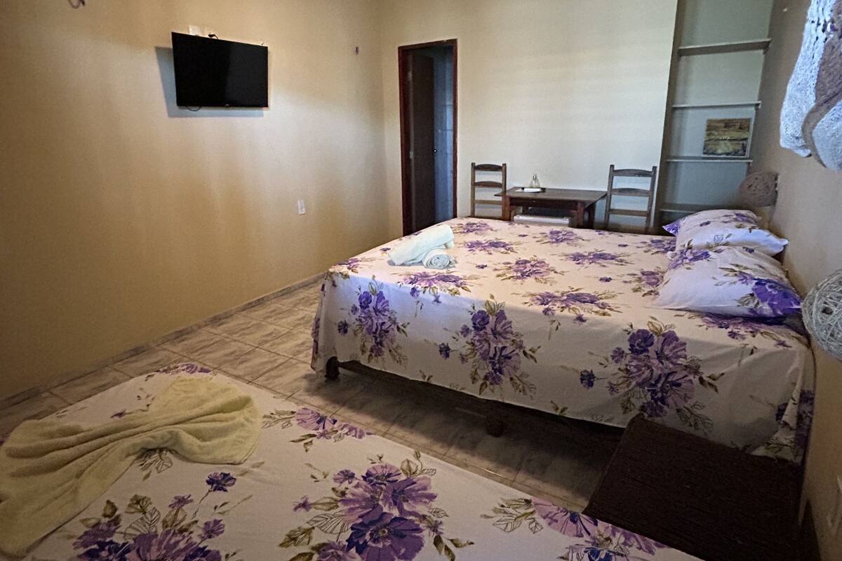 Quarto com duas camas, ventilador de teto, uma televisão pequena e porta que dá para outro cômodo.
