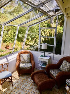 Relax in the serene conservatory with a lush garden view, featuring a wicker armchair, a small table, and a single chair.