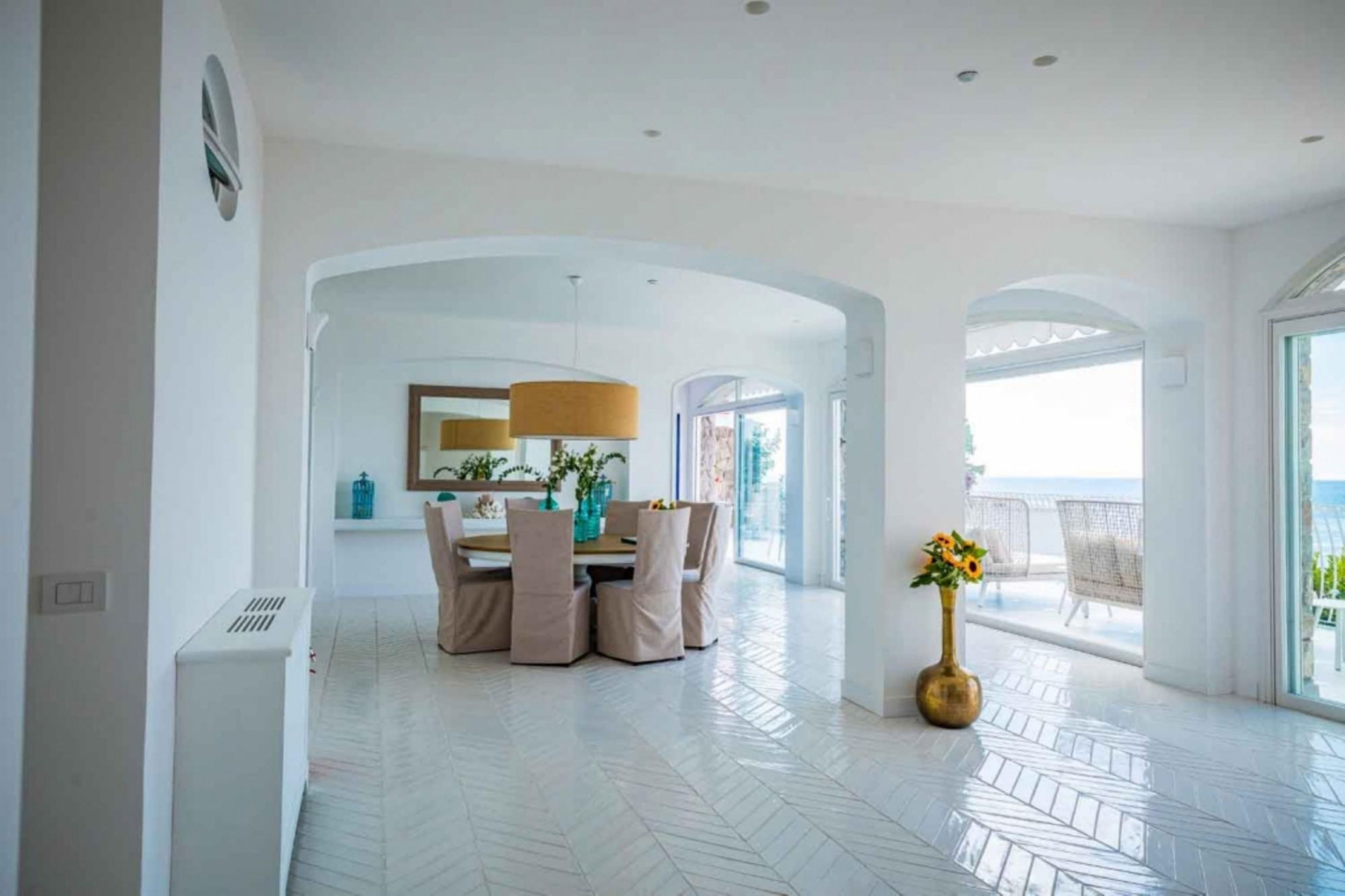 Um quarto espaçoso com piso em padrão herringbone, uma área de jantar com mesa e cadeiras, e uma grande janela com vista para o mar.
