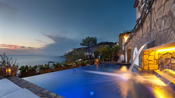 A swimming pool with a waterfall feature, stone walls, and a view of the sea at dusk. - Villa dei Sogni in Massa Lubrense (Massa Lubrense)