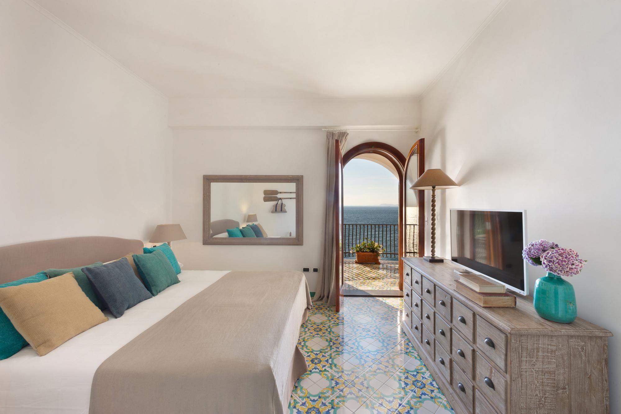Un dormitorio con cama, cómoda, televisor, espejo y vista al mar.