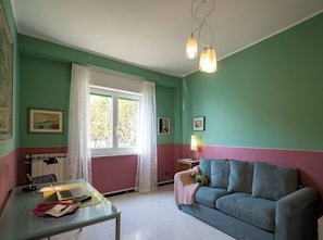 A room with a green and pink wall color scheme, a glass table with a laptop, a blue sofa, and a window with curtains.