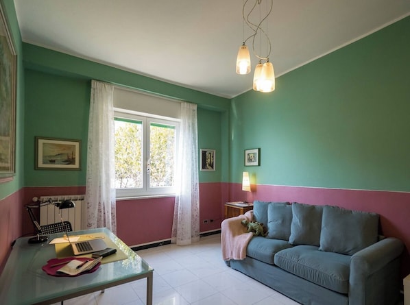 A room with a green and pink wall color scheme, a glass table with a laptop, a blue sofa, and a window with curtains.