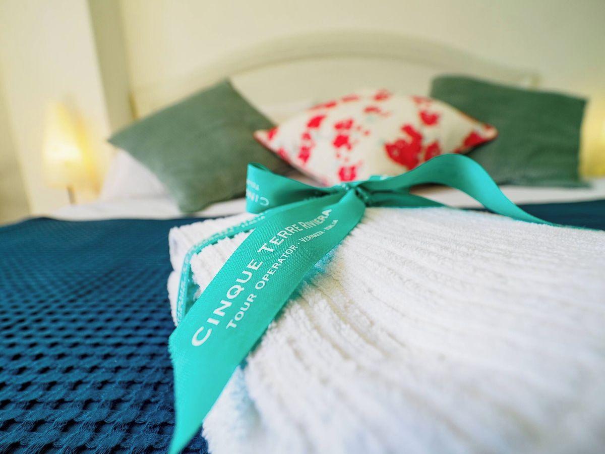 Un letto rifatto con cura, con un nastro color petrolio legato ai cuscini, una coperta bianca e una trapunta blu con texture.