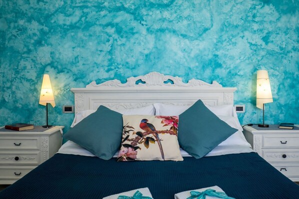 Quarto com parede verde-água texturizada, cabeceira branca ornamentada, duas mesas de cabeceira com abajures, e cama com colcha azul-escuro e almofadas florais.