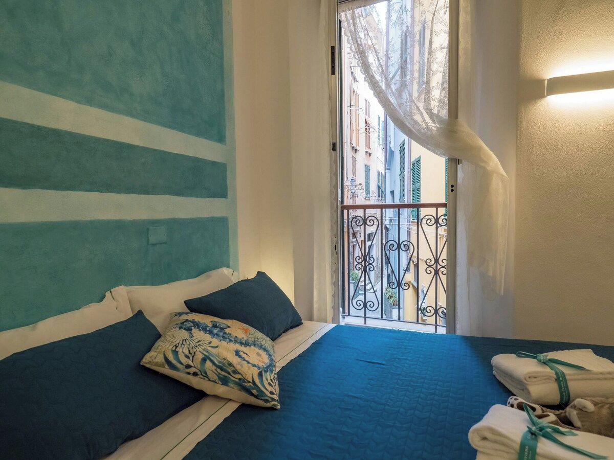 Ein Schlafzimmer mit Bett, Kissen, einem Fenster mit Blick auf Gebäude und einem Balkon mit Geländer.