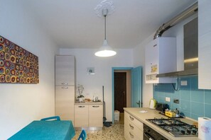 Une cuisine compacte équipée d’une cuisinière à gaz, d’un four à micro-ondes et d’un évier. On y trouve une table à manger avec des chaises, un luminaire au plafond et une porte donnant sur une autre pièce.
