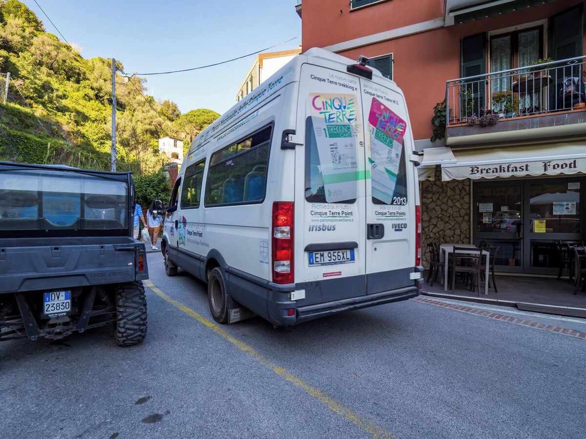 Ein weißer Van mit der Aufschrift 'Cinq Terre Trekking' und 'Cinq Terre Point' auf der Rückseite, geparkt in einer Straße vor einem Gebäude, das einen Balkon und ein Schild mit der Aufschrift 'Frühstücksessen' hat.