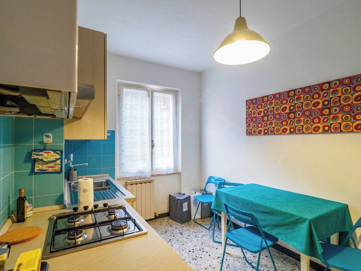 Une cuisine de taille réduite, équipée d’une cuisinière à gaz, d’une fenêtre avec des rideaux, d’une table à manger avec des chaises, et ornée d’un tableau abstrait aux couleurs vives.
