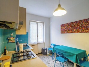 A compact kitchen with a gas stove, a window with curtains, a dining table with chairs, and a colorful abstract painting on the wall.