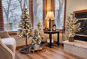 Una sala de estar acogedora con chimenea, una mesita de noche con lámpara y dos árboles de Navidad artificiales.